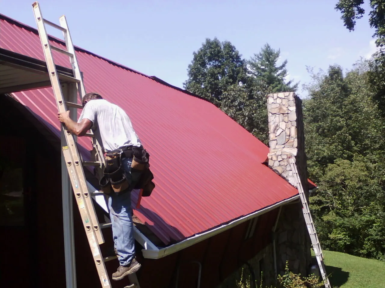 Roofer on a ladder beside a red metal roof and stone chimney — Soffit & Fascia Repair by Roofing Brothers in Piney Flats